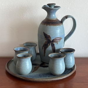 May include: A set of six blue and brown ceramic cups and a matching jug with a butterfly design. The cups and jug are on a round, blue and brown ceramic tray.