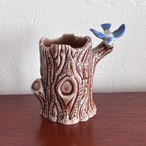 May include: A brown ceramic tree stump-shaped vase with a blue bird perched on a branch. The vase has a textured bark design and a hollowed-out interior. The bird has blue wings and a white chest.