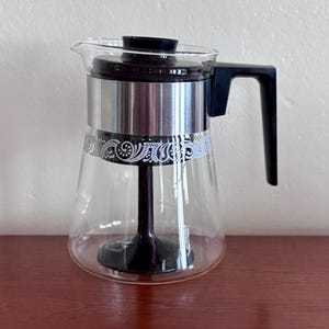 May include: A clear glass coffee carafe featuring a black plastic lid and handle, a central black plastic stem, and a silver metal band around the upper section. The glass body is decorated with a white filigree pattern around the middle.