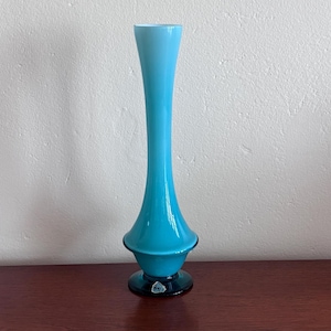 May include: A tall, slender glass vase featuring a vibrant turquoise blue body that transitions to a lighter sky blue at the flared rim. The vase has a bulbous midsection and a dark, circular base with a small, clear sticker label attached to it.