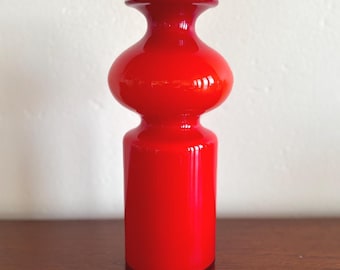 May include: A bright red glass vase with a cylindrical base, a rounded midsection, and a flared rim. The glass has a smooth, glossy finish that reflects light, and the top edge of the rim is white. The vase stands upright with a symmetrical shape.