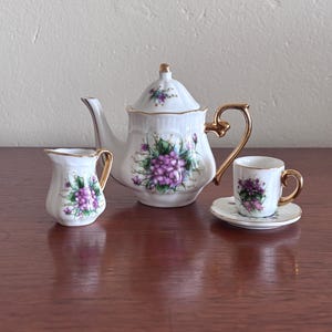 Könnte beinhalten: Ein Miniatur-Teeservice aus Porzellan bestehend aus einer Teekanne, einem Milchkännchen sowie einer Tasse mit Untertasse. Jedes Teil ist weiß mit einem Blumenmuster aus violetten Blüten und grünen Blättern sowie goldenen Rändern und Griffen.