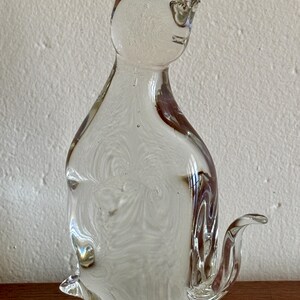 May include: A clear glass cat figurine with a seated pose. The sculpture has a swirled, textured interior and brown accents. The cat has pointed ears and a curved tail. The figurine is displayed on a wooden surface.