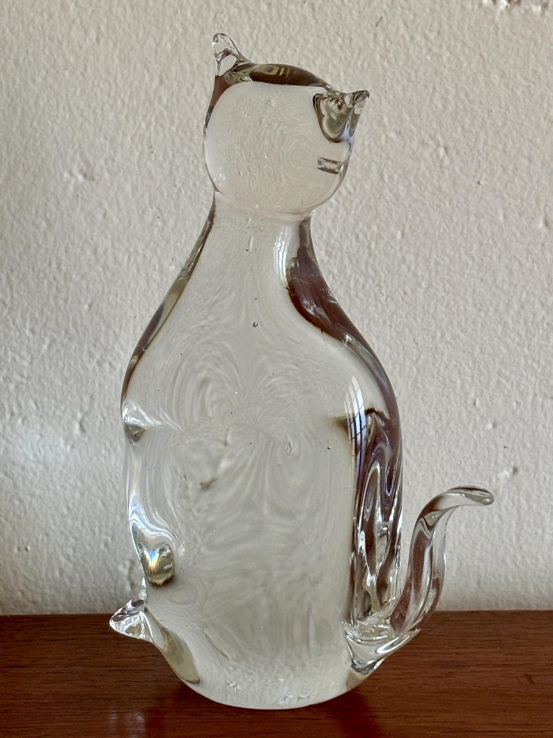 May include: A clear glass cat figurine with a seated pose. The sculpture has a swirled, textured interior and brown accents. The cat has pointed ears and a curved tail. The figurine is displayed on a wooden surface.