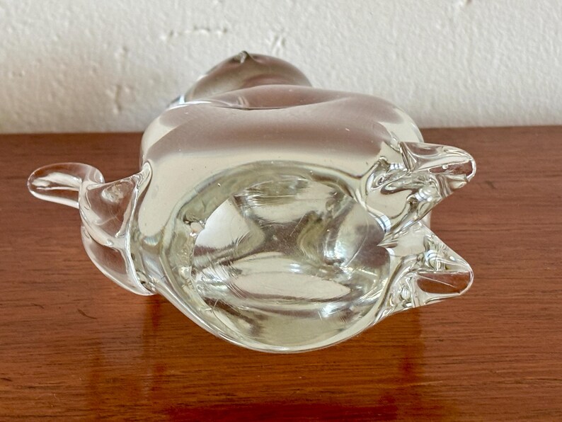 May include: A clear glass ashtray with a unique, organic shape. The ashtray has a rounded body with a flared rim and two curved extensions. The glass is transparent, allowing light to pass through, and sits on a wooden surface.