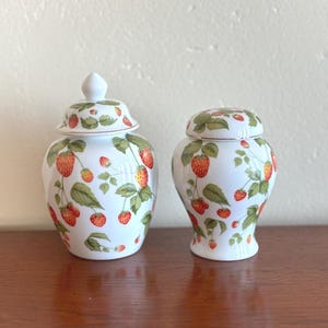 May include: Two white ceramic jars with lids, decorated with a strawberry pattern. The jars display red strawberries, green leaves, and white backgrounds. The jars are on a wooden surface.