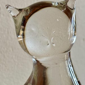 May include: A clear glass owl figurine with a rounded body and pointed ears. The owl's face is a lighter shade of clear glass, and the body has a slight brown tint. The figurine is a decorative item.