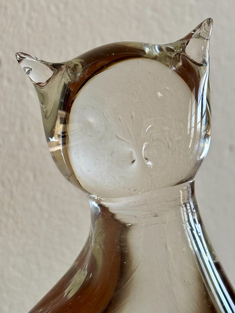 May include: A clear glass owl figurine with a rounded body and pointed ears. The owl's face is a lighter shade of clear glass, and the body has a slight brown tint. The figurine is a decorative item.