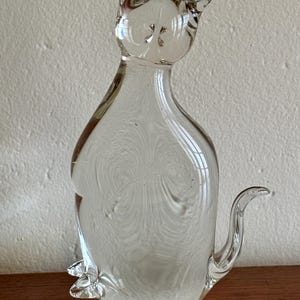 May include: A clear glass cat figurine with pointed ears and a curled tail. The transparent sculpture has a smooth, rounded body and sits upright. The cat's features are subtly defined, creating a minimalist aesthetic. The figurine is a decorative item.