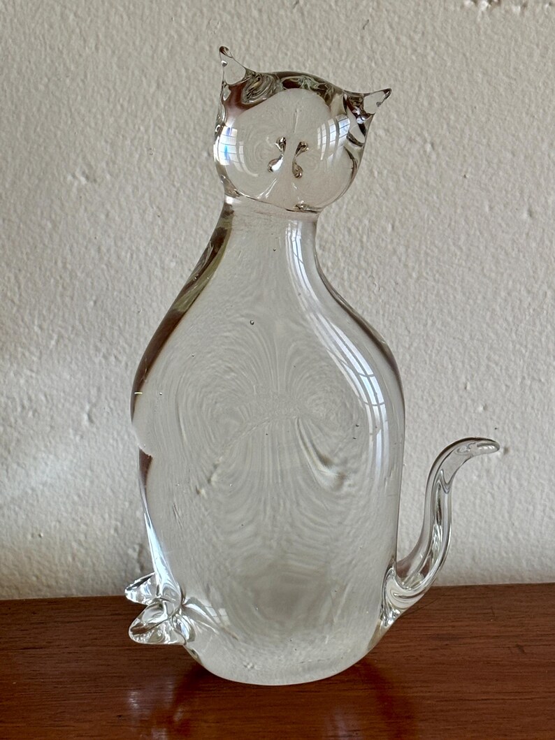 May include: A clear glass cat figurine with pointed ears and a curled tail. The transparent sculpture has a smooth, rounded body and sits upright. The cat's features are subtly defined, creating a minimalist aesthetic. The figurine is a decorative item.