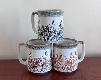 May include: Three white ceramic mugs with gray rims and handles are stacked on a wooden surface. Each mug features a different botanical design in black, brown, and orange. The mugs are approximately 4 inches tall.