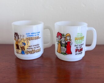 May include: Two white ceramic mugs with humorous cartoon illustrations and text. One mug features a woman, child, and text: "JUST WHEN I HAD LEARNED ALL THE ANSWERS... THEY STARTED ASKING ALL NEW QUESTIONS!" The other mug has a cartoon and text: "A FOOL AND HIS MONEY MAY BE SOON PARTED BUT THINK OF ALL THE FUN HE HAS!!"