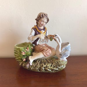 May include: A porcelain figurine featuring a child with brown hair wearing a white shirt, purple vest, and tan trousers, sitting on a textured green base. The child is feeding two white geese with grey-tipped wings, next to a small green planter.