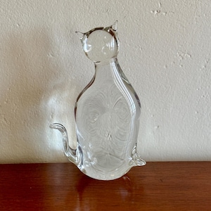 May include: A clear glass cat figurine with pointed ears and a curled tail. The cat is sitting upright, with a detailed body and a smooth, transparent finish. The figurine is displayed on a wooden surface.