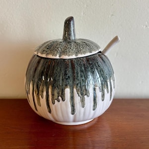 May include: A ceramic sugar bowl with a lid and spoon. The bowl is round with a white base and a dark green and brown dripping glaze. The lid has a textured pattern and a small handle. The spoon is light beige.