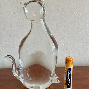 May include: A clear glass cat figurine with pointed ears and a curved tail. The cat is next to a ChapStick lip balm tube with a mango design. The figurine is on a wooden surface.