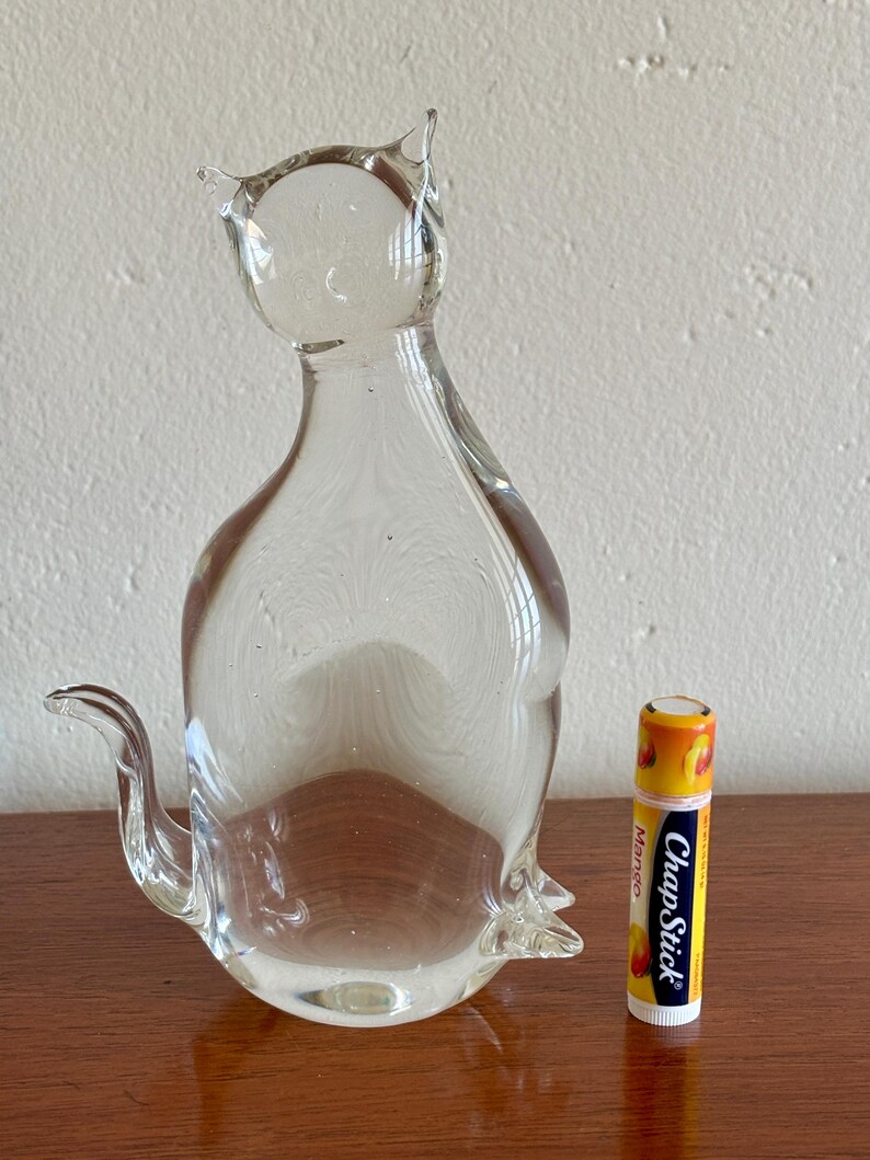 May include: A clear glass cat figurine with pointed ears and a curved tail. The cat is next to a ChapStick lip balm tube with a mango design. The figurine is on a wooden surface.