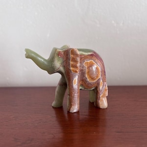 May include: A small, carved stone elephant figurine. The elephant is primarily green, with brown and reddish-brown marbling on its body. The trunk is raised, and the figurine has a smooth, polished finish. A decorative item.