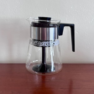 May include: A clear glass coffee percolator with a black handle and lid. The percolator has a silver band with a white floral design. The coffee maker is sitting on a brown surface.