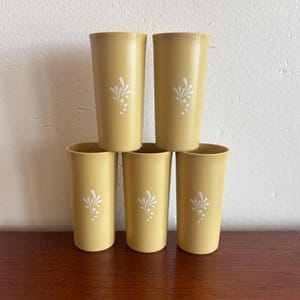 Könnte beinhalten: Ein Satz von fünf gelben Vintage-Trinkgläsern. Jedes Trinkglas hat ein weißes Blumendesign. Die Trinkgläser sind auf einer Holzoberfläche vor einer weißen Wand gestapelt.