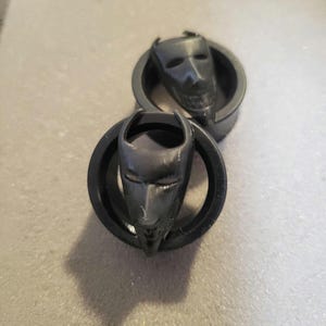 May include: Two black devil face plugs. Each plug features a detailed devil face with horns, eyes, and a mouth. The plugs are set in a circular frame. The plugs are made of a dark material.