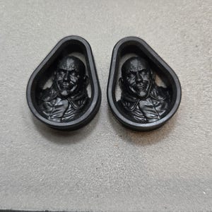 May include: Two black, teardrop-shaped ear gauges with a detailed relief of a smiling man's face. The gauges are made of a solid, dark material and are displayed against a neutral background.
