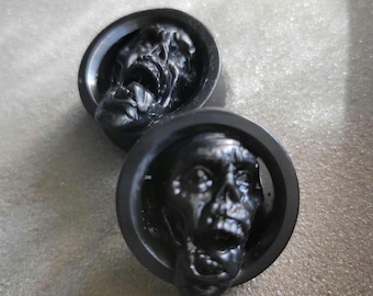 May include: Two black, circular plugs with a zombie skull design. The skulls have open mouths and detailed features. The plugs are set against a textured, light gray background.