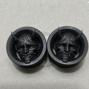May include: Two black ear gauges featuring a detailed, carved oni mask design. The masks have horns, a wide grin, and intricate facial features. The gauges are circular with a raised rim.