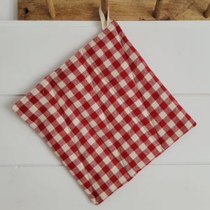 Op de afbeelding: Een rood-wit geruite, gewatteerde pannenlap met een ophanglus. De vierkante pannenlap hangt aan een houten kapstok. Het patroon is een klassiek ruitjesontwerp.