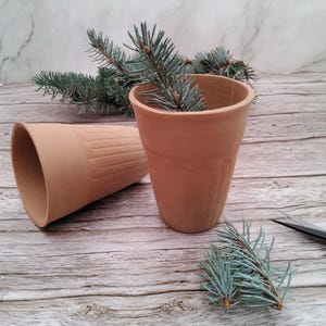 Könnte beinhalten: Zwei kleine, terrakottafarbene Tontöpfe, einer aufrecht und der andere auf der Seite, mit strukturierten vertikalen Linien. Ein Zweig blau-grüner Nadeln befindet sich in einem Topf. Der Hintergrund umfasst eine Holzoberfläche und eine marmormuster Wand.