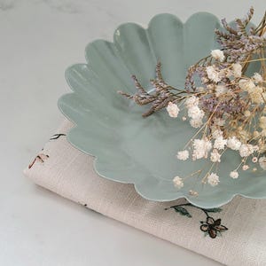 Peut inclure: Un plat en céramique de couleur vert clair à bords festonnés avec des fleurs blanches séchées dessus. Le plat est posé sur un tissu beige à motif floral.