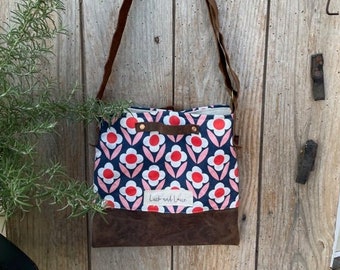 May include: A blue and pink floral print crossbody bag with a brown leather strap and bottom. The bag has a brown leather flap with the text "Luck and Love" in white.