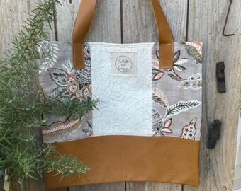 May include: A brown leather tote bag with a patchwork design. The bag has a white fabric panel with a floral pattern and a brown leather bottom. The bag has two brown leather straps.