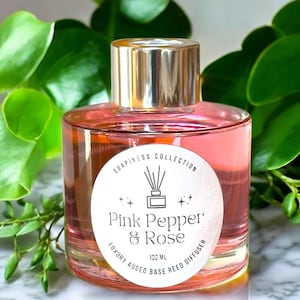 May include: A clear glass bottle with a silver lid filled with a pink liquid. The bottle has a white label with the text "Soapiness Collection Pink Pepper & Rose Luxury Augeo Base Reed Diffuser 100 ML".
