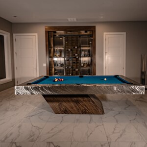 May include: A blue felt pool table with a brushed metal frame and a wooden base. The table is set up in a game room with a wine cellar in the background.