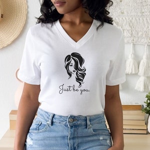 May include: White v-neck t-shirt with a black graphic of a woman's face and the text "Just be you."