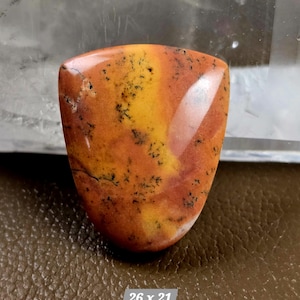 May include: A polished, shield-shaped gemstone with a blend of orange, yellow, and red hues. The stone features dark, dendritic inclusions, and the dimensions are 26 x 21.