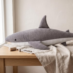 May include: A plush shark toy crafted from grey corduroy fabric with a white underbelly. The shark features a prominent dorsal fin, side fins, and a tail fin. It has a small black button eye on the side of its head and stitched gill details.