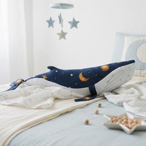 May include: A navy blue whale-shaped pillow with a celestial design of stars, moons, and suns, resting on a bed with a white blanket. A star-shaped bowl with wooden beads and a pillow with a crescent moon design are also visible.