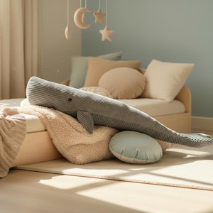 K&ouml;nnte beinhalten: Ein graues Wal-Pl&uuml;schtier aus Cord liegt auf einem Bett mit beigen und blauen Kissen. Das Zimmer verf&uuml;gt &uuml;ber einen hellen Holzrahmen und einen cremefarbenen Teppich. Ein Mobile mit Mond- und Sternformen h&auml;ngt &uuml;ber dem Bett.