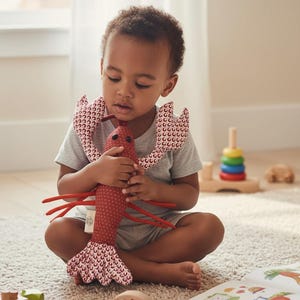 May include: A child sits on a carpet, holding a red and white polka-dotted lobster plush toy. The toy has red legs and a patterned collar. Wooden toys and a colorful book are nearby.