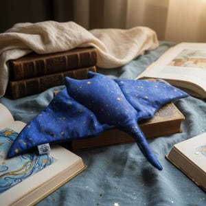 K&ouml;nnte beinhalten: Ein blaues Pl&uuml;sch-Stachelrochen-Spielzeug mit goldenen Sternen liegt auf einem alten Buch. Das Spielzeug liegt auf einer blauen Stoffoberfl&auml;che, umgeben von offenen B&uuml;chern mit Illustrationen. Ein Stapel alter B&uuml;cher und ein wei&szlig;es Tuch befinden sich im Hintergrund.