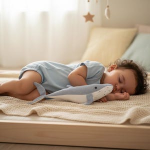 Peut inclure: Un bébé endormi, vêtu d'un body bleu clair, est allongé sur un lit avec une couverture beige. Il serre contre lui un jouet en peluche baleine bleu clair à pois blancs. La chambre a une palette de couleurs neutres.