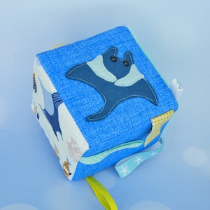 Può includere: Un cubo di tessuto blu e bianco con un design a manta. Il cubo presenta una varietà di trame e motivi, tra cui un motivo a quadri, un motivo a pois e un motivo a stelle. Il cubo ha un anello giallo attaccato.