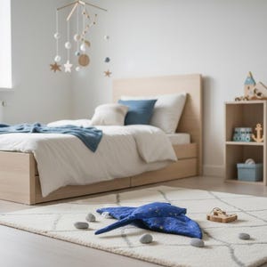 K&ouml;nnte beinhalten: Ein Kinderzimmer mit einem hellen Holzbett, wei&szlig;er Bettw&auml;sche und blauen Akzenten. Ein blaues Kissen in Form eines Mantarochens und Holzspielzeug liegen auf dem Teppich. Ein Mobile h&auml;ngt &uuml;ber dem Bett.