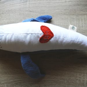 Peut inclure: Une baleine en peluche blanche avec des nageoires et une queue bleues. Un c&oelig;ur rouge avec le mot "Love" cousu sur le c&ocirc;t&eacute;. La baleine est pos&eacute;e sur une surface en bois.