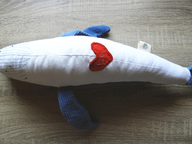 Peut inclure: Une baleine en peluche blanche avec des nageoires et une queue bleues. Un c&oelig;ur rouge avec le mot "Love" cousu sur le c&ocirc;t&eacute;. La baleine est pos&eacute;e sur une surface en bois.