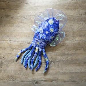 Puede incluir: Un juguete de pulpo de peluche con un dise&ntilde;o de noche estrellada azul y blanco, con largos tent&aacute;culos azules. El juguete tiene un volante transl&uacute;cido e iridiscente alrededor de la cabeza, sobre una superficie de madera.