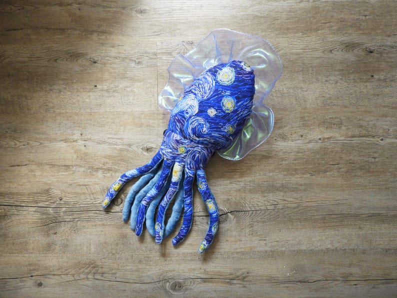 Puede incluir: Un juguete de pulpo de peluche con un dise&ntilde;o de noche estrellada azul y blanco, con largos tent&aacute;culos azules. El juguete tiene un volante transl&uacute;cido e iridiscente alrededor de la cabeza, sobre una superficie de madera.