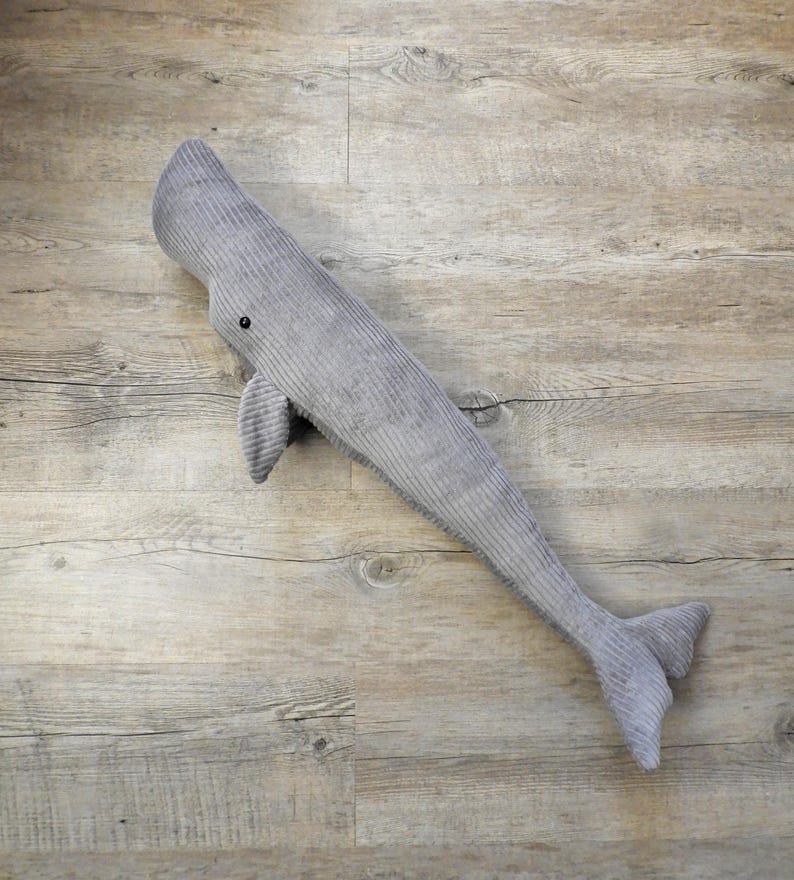 K&ouml;nnte beinhalten: Ein graues Wal-Pl&uuml;schtier aus Cord mit einem kleinen schwarzen Auge und Flosse. Der Wal liegt auf einer Holzoberfl&auml;che. Das Pl&uuml;schtier ist etwa 76 cm lang.